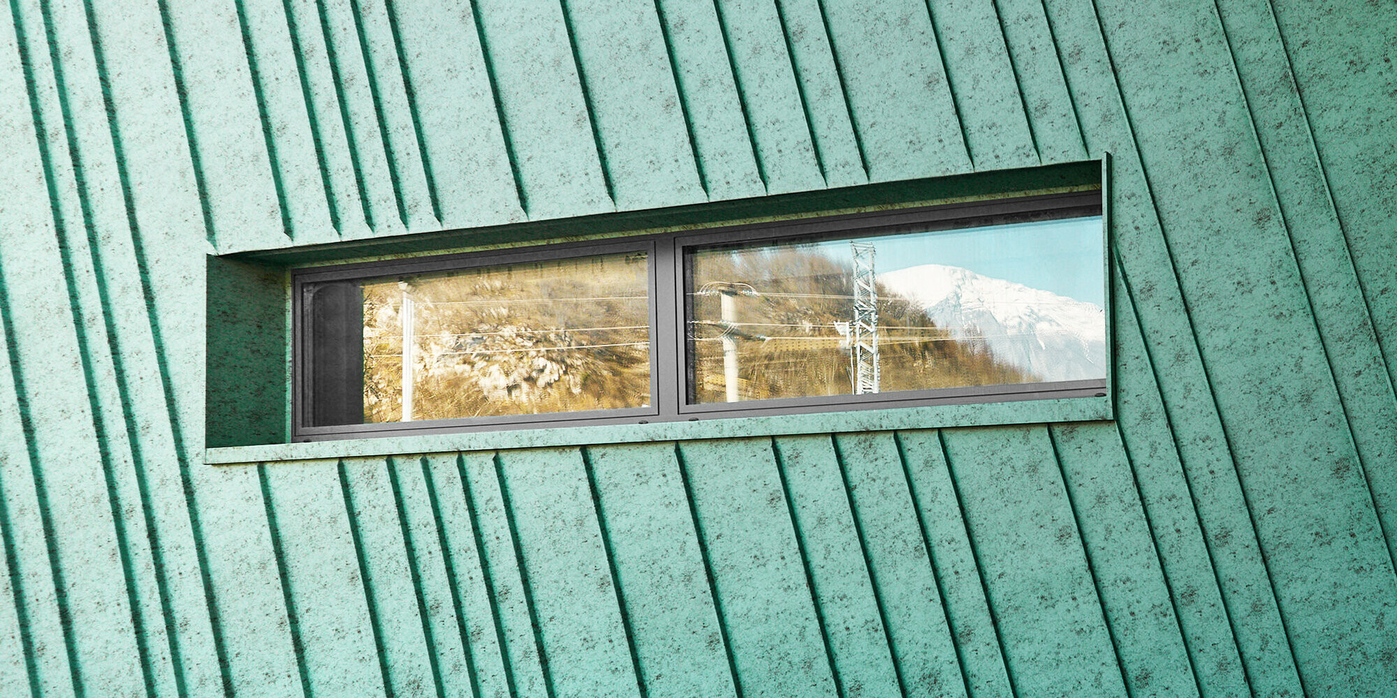 Detail zblízka mírně zkosené fasády s malým, podlouhlým oknem odrážejícím hory a elektrické kabely. Boční pohled na fasádu a prosklené průčelí se vstupem.