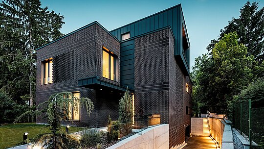 Fotografie ukazuje moderní, jasně strukturovaný exteriér vícegenerační vily v Budapešti za soumraku. Tmavě červenohnědé cihlové fasády se setkávají s precizně provedeným hliníkovým obkladem PREFALZ v antracitové barvě P.10, jehož svislé linie vytvářejí silný architektonický akcent. Teple osvětlená okna dodávají budově hloubku a atmosféru. Jemně osvětlená příjezdová cesta do podzemních garáží elegantně vede podél budovy a zdůrazňuje vysoce kvalitní a povětrnostním vlivům odolnou kombinaci cihel a hliníku PREFA.