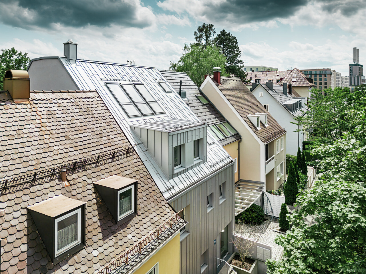 Pohled na střechy řadové zástavby rodinných domů v kombinací s tradičními taškovými střechami a moderních hliníkových střech Prefalz ve stříbrné metalíze. Jeden dům vyniká svou zrekonstruovanou, lesklou kovovou střechou a odpovídajícími střešními okny. Zelená, hustá vegetace zahrad dotváří obraz a vytváří přirozený kontrast k městské architektuře. V pozadí jsou vidět obrysy moderních městských budov.
