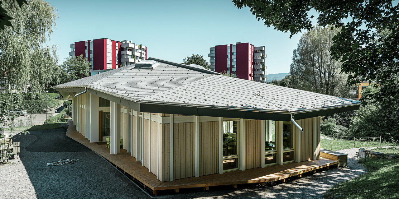Jednopodlažní školka s vertikálními fasádními lamely a hliníkovou střechou od PREFA (falcované šablony 44 x 44) v barvě P.10 světle šedé
