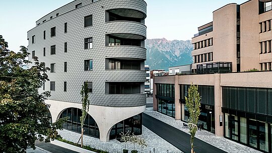 Pohled na Bernerhaus v Buchsu s jeho výrazným trojúhelníkovým tvarem a jemně zaoblenými balkony na pozadí alpských hor. Souvislá fasáda z PREFA fasádních šindelů v barvě P.10 světle šedé obaluje budovu jako šupinovitý hliníkový plášť a vytváří živou hru světla a stínu. Nad masivním betonovým základem s elegantními arkádami působí lehká, povětrnostním vlivům odolná hliníková fasáda PREFA obzvláště precizně, trvanlivě a nenáročně na údržbu – v městském prostředí architektonicky poutavá.