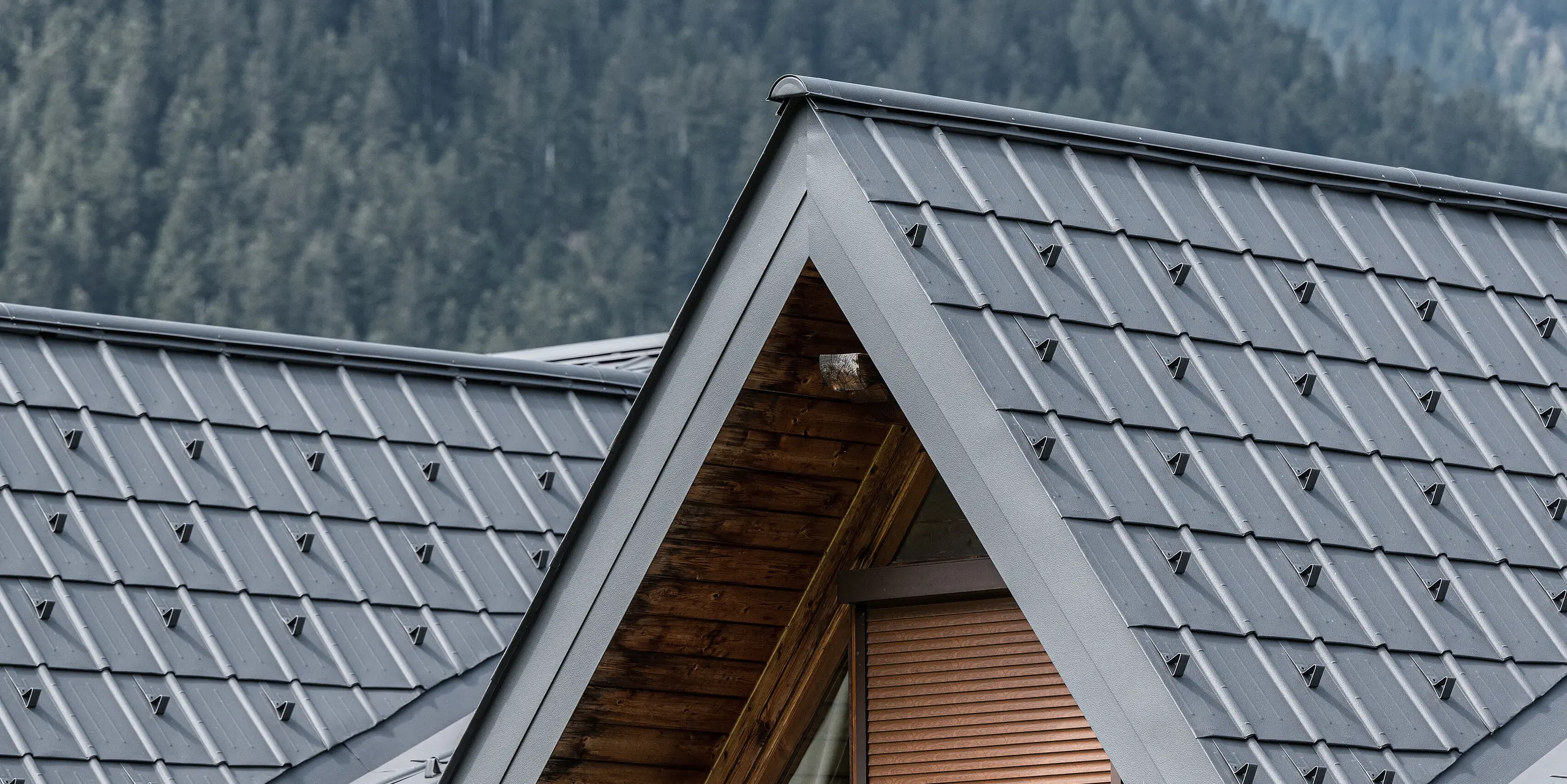 Detailní záběr na vikýř na alpském domě v Auronzo di Cadore, vybavený falcovanými taškami PREFA ve světle šedé barvě. Jemně strukturované hliníkové panely krásně kontrastují s přírodním dřevem střešní konstrukce a nabízejí vysoce kvalitní ochranu před alpským počasím a zároveň se hladce začleňují do horské lesní krajiny.