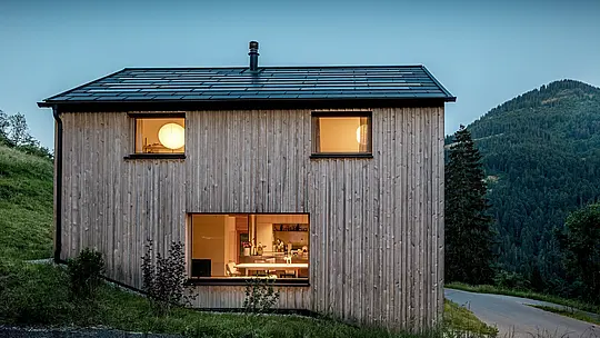 Minimalistický dřevěný dům Immi v Ebnitu září za soumraku na zalesněném horském svahu. Na čistých liniích sedlové střechy se černé solární střešní panely PREFA téměř neviditelně prolínají se střešním panelem R.16 v černé barvě P.10., který generuje solární energii, poskytuje budově trvalou ochranu a podtrhuje klidnou, moderní architekturu tohoto alpského dřevěného domu.