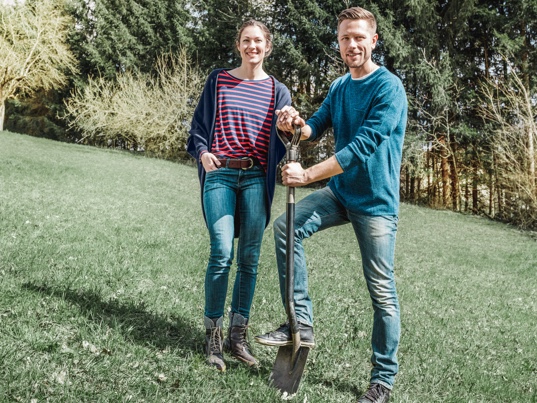 Mladý páreček stojí na svém ještě nezastavěném stavebním pozemku. Muž drží v ruce rýč. V této stavební fázi si stavebníci opatřují nabídky na stavbu domu.