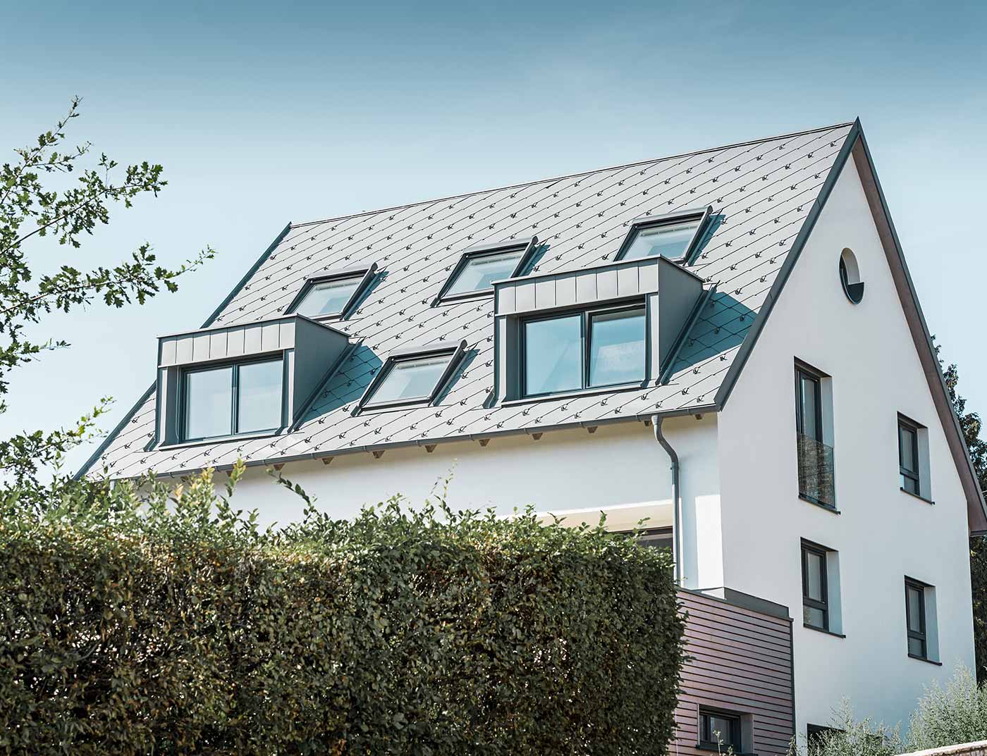 PREFA střešní šablona 44 × 44, barva světle šedá - reference Klasická sedlová střecha se dvěma vikýři a několika střešními okny, pokrytá PREFA střešními šablonami 44 × 44 světle šedé barvy.