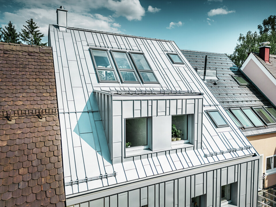 Moderní řadový dům po rekonstrukci střechy a fasády s hliníkovými výrobky PREFA. Střechu tvoří hliníkové pásy v barvě stříbrné metalízy, které mají texturovaný, matný povrch a odrážejí světlo. Fasáda domu je také obložena vertikálními hliníkovými prvky, které poskytují kontrast k tradičním, terakotově zbarveným taškovým střechám přilehlých domů.